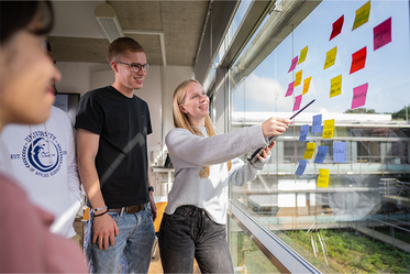 Studentin erklärt anderen Studierenden Post-Its die an einer Fensterscheibe befestigt sind
